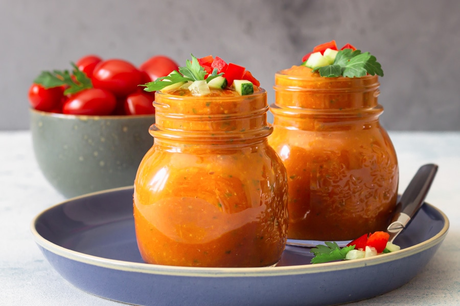 Przepis na gazpacho Obrazek nastroju: Dwa słoiki z zakrętką wypełnione czerwonym gazpacho ustawione na niebieskim talerzu, w tle miska pomidorów